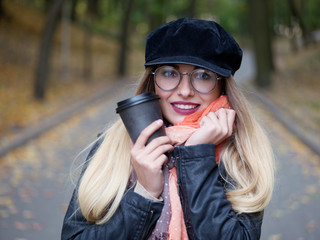 Portrait of a beautiful stylish young blonde girl with makeup in round glasses of a leather jacket, cap. Frozen girl drinks hot drink in a black mug in the autumn cold park