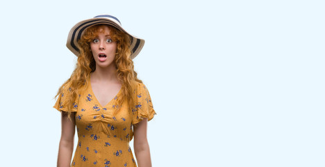 Young redhead woman wearing summer hat and dress scared in shock with a surprise face, afraid and...