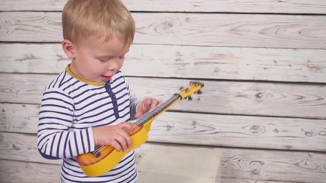 happy one year old boy plays his guitar or ukulele and sings songs, sitting in the wooden room.