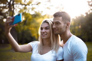 Cute loving couple standing on grass in nature green park with beautiful sundown light using mobile phone take a selfie.