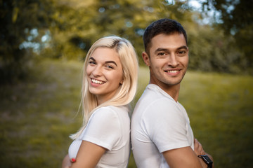 Fototapeta premium Cute loving couple standing back together on grass in nature green park with beautiful sundown light. Looking camera.