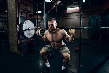 Bearded tattoed guy with six-pack squating and lifting barbell in crossfit gym. Weight plates in...
