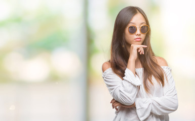 Fototapeta premium Young asian woman wearing sunglasses over isolated background with hand on chin thinking about question, pensive expression. Smiling with thoughtful face. Doubt concept.