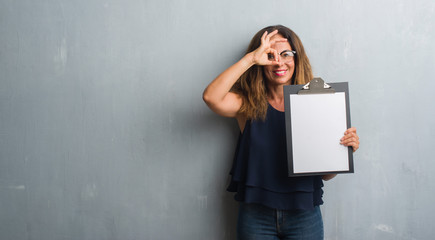 Middle age hispanic woman standing over grey grunge wall holding clipboard with happy face smiling doing ok sign with hand on eye looking through fingers