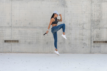 Fototapeta premium Beautiful caucasian athletic girl with long black hair wearing blue jeans and black sports bra and wearing a cap jumps in a concrete stadium on a bright sunny day