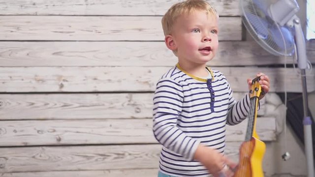 happy one year old boy plays his guitar or ukulele and sings songs, sitting in the wooden room.