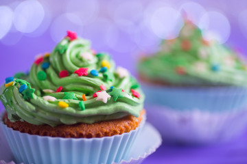 Closeup two New Year cupcakes with creamy green top with colorful stars and sprinkles on purple bokeh background.