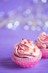 Closeup two cupcakes with creamy pink and white top decorated with little hearts on purple bokeh background.