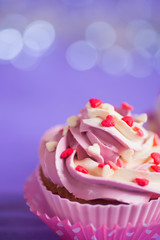 Closeup cupcake with creamy pink and white top decorated with little hearts on purple bokeh background.