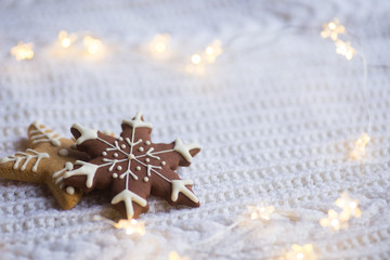 Christmas glazed cookie in form of star and snowflake with warm garland lights on white knitted background.