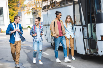 Obraz premium smiling multicultural friends with backpacks walking near travel bus at street