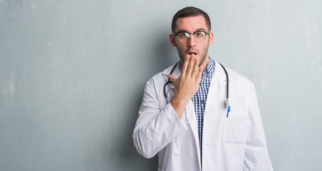 Young caucasian man over grey grunge wall wearing doctor uniform cover mouth with hand shocked with shame for mistake, expression of fear, scared in silence, secret concept
