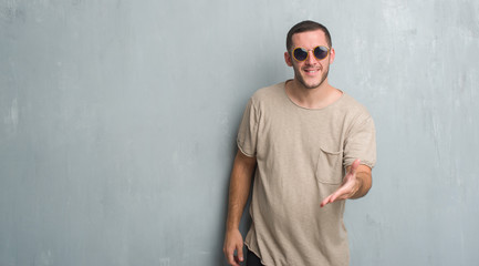 Obraz premium Young caucasian man over grey grunge wall wearing sunglasses smiling friendly offering handshake as greeting and welcoming. Successful business.