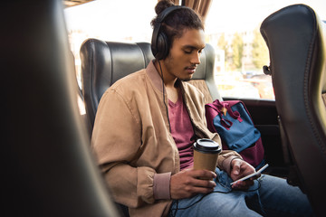 multiracial male tourist in headphones sitting with paper coffee cup and using smartphone in bus