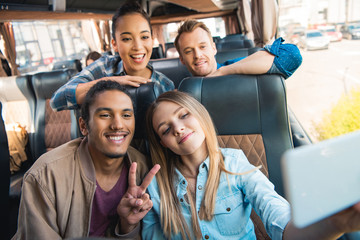 young mixed race man doing peace sign with friends taking selfie on smartphone during trip on...