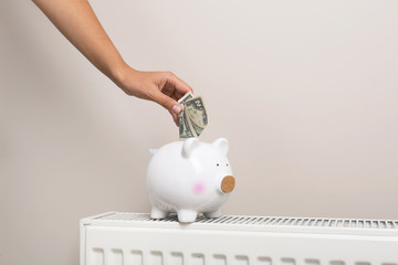 Woman putting money into piggy bank on heating radiator against light background