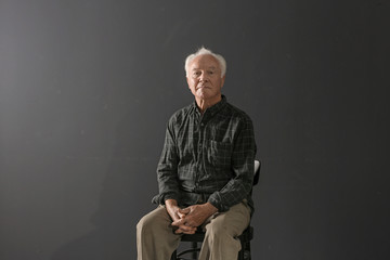 Portrait of poor elderly man sitting on chair against dark background - Powered by Adobe