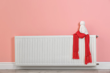 Heating radiator with knitted cap and scarf near color wall. Space for text