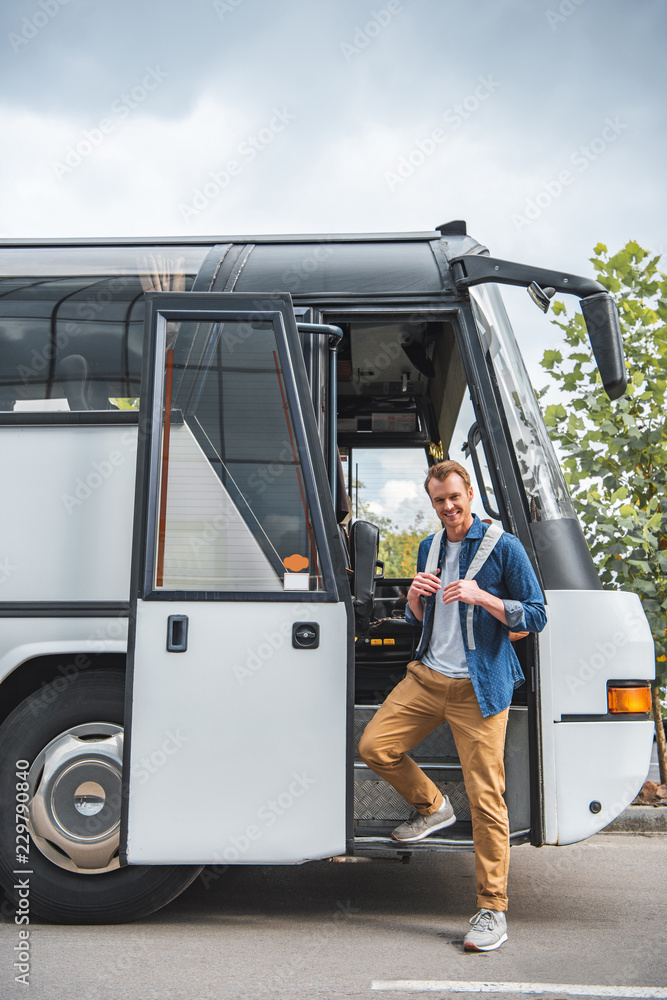 Wall mural smiling man with backpack posing near travel bus at urban street