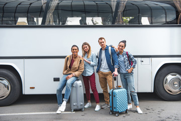 young multicultural friends with wheeled bags posing near travel bus at city street