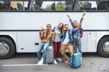 cheerful multiethnic tourists with travel bags doing peace signs near bus at street
