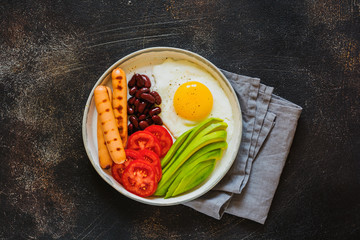 Traditional English breakfast with fried eggs, avocado, sausages grill, beans, and tomatoes on dark concrete background. Top view.