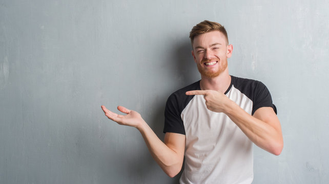 Young redhead man over grey grunge wall amazed and smiling to the camera while presenting with hand and pointing with finger.