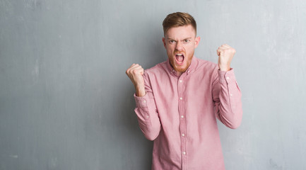 Young redhead man over grey grunge wall wearing pink shirt angry and mad raising fist frustrated and furious while shouting with anger. Rage and aggressive concept.