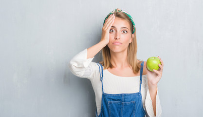 Beautiful young woman over grunge grey wall eating green apple stressed with hand on head, shocked with shame and surprise face, angry and frustrated. Fear and upset for mistake.