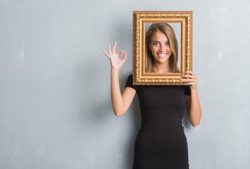 Beautiful young woman over grunge grey wall holding vintage frame doing ok sign with fingers,...