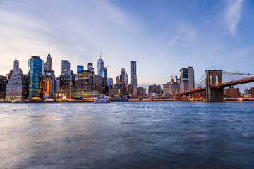 Obraz na płótnie Canvas Outdooors view on NYC New York City Brooklyn Bridge Park by east river, cityscape skyline at sunset, dusk, twilight, blue hour, dark night, skyscrapers, buildings, waves, blurred, blurry tour boat