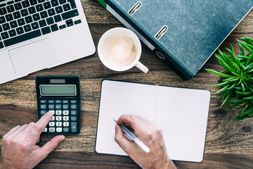 top view of person at desk using calculator while taking notes
