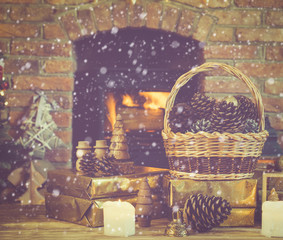 Christmas composition, pine cones in wicker basket, wooden ornaments, golden presents on the table in front of fireplace with woodburner, candles and garlands, selective focus, snow, toned