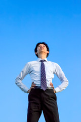 青空をバックに腰に両手を当て見上げる若いYシャツ姿の男性1人立ち姿。ビジネス、成功、野心、向上心イメージ