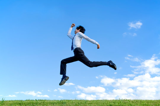 青空をバックにジャンプするYシャツ姿の若いビジネスマン1人。元気・パワー・喜び・挑戦イメージ