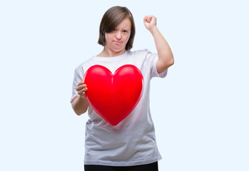 Young adult woman with down syndrome holding red heart over isolated background annoyed and frustrated shouting with anger, crazy and yelling with raised hand, anger concept