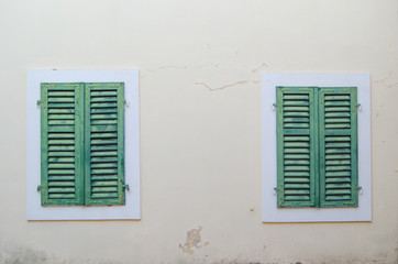 Two Windows are closed by green shutters against a beige wall.