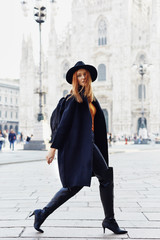 Fashion model walking by the Duomo of Milan looking into the camera wearing a hat and a dark coat...