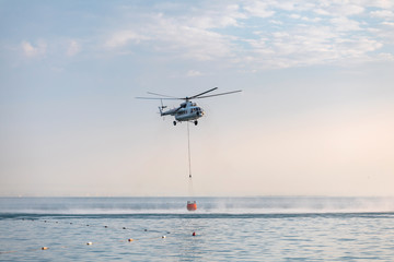 A helicopter with a red basket descends over the sea to scoop water against the background of the dawn orange sky and the silhouette of the city in the distance