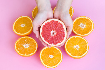Female hands holding half of a juicy ripe red grapefruit above oranges cut in half, top view.