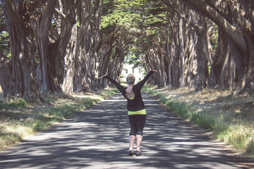 Blonde female raises arms up at the Cypress Tree Tunnel on Point Reyes National Seashore in Marin County in Northern California