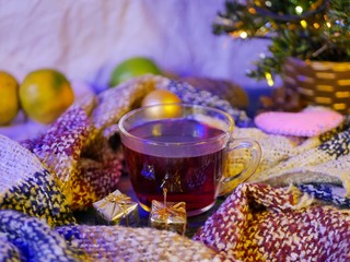 A cup of tea wrapped in a fluffy scarf against the backdrop of a Christmas tree and Christmas tree decorations, golden gifts, evening lighting, the concept of Christmas, home winter seasonal holidays