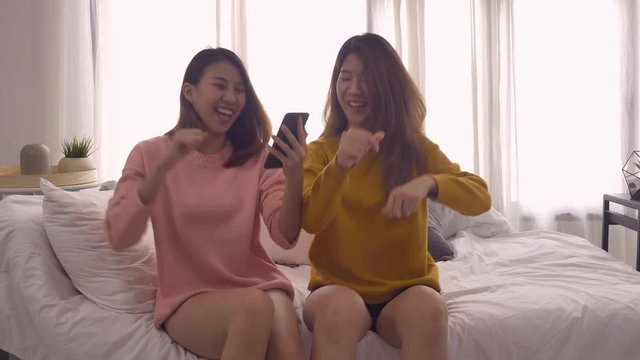 Happy Asian lesbian lgbt couple enjoy entertainment in living room. Beautiful women lying on a sofa listen to music on smartphone at home.