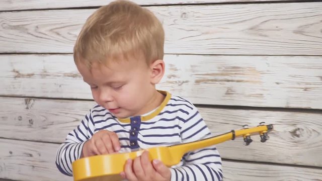 One year old boy plays his guitar or ukulele and sings songs, sitting in the wooden room.