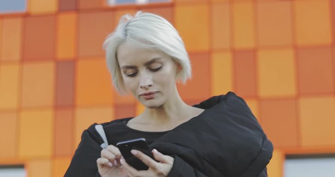 Half length portrait of a beautiful smiling blone woman with short hair wearing jacket coat chatting online by a mobile phone on street background. 4k video evening urban city style