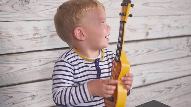 One year old boy plays his guitar or ukulele and sings songs, sitting in the wooden room.