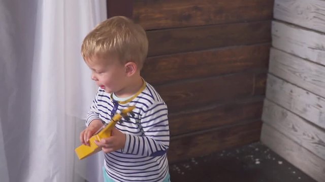 One year old boy plays his guitar or ukulele and sings songs, sitting in the wooden room.