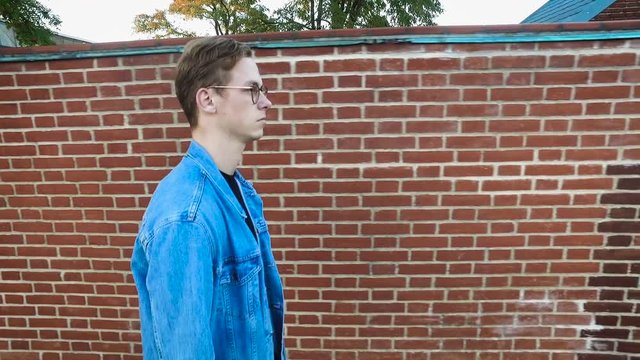 Upset sad hipster guy with glasses walking in the town by the brick wall and thinking about his next steps in his life