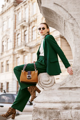 Outdoor full body fashion portrait of  fashionable woman wearing sunglasses, white turtleneck, green suit, blazer, trousers, leopard print boots, holding suede bag, posing in street of european city