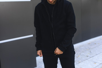 Young man zipping up his hoodie. Man dressing black sport clothes. Grey urban background. 